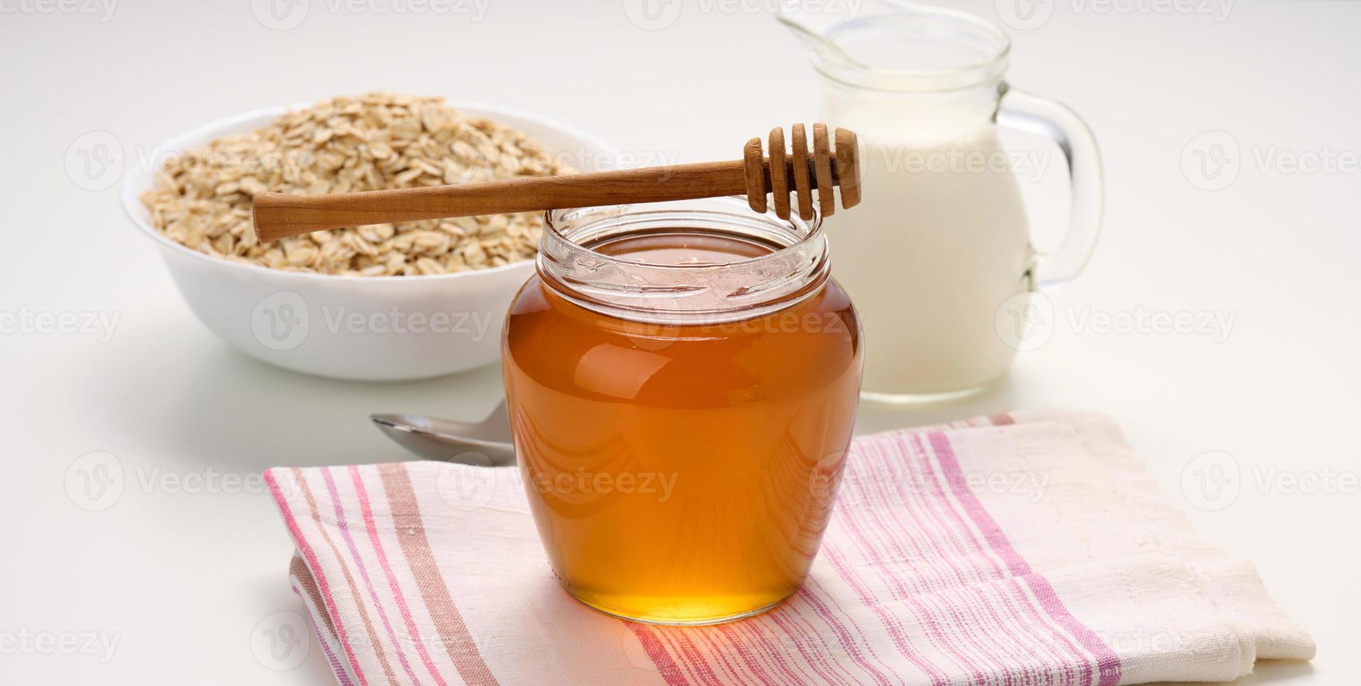 honey in a glass transparent jar and a wooden stick on a white table, behind a decanter with milk photo