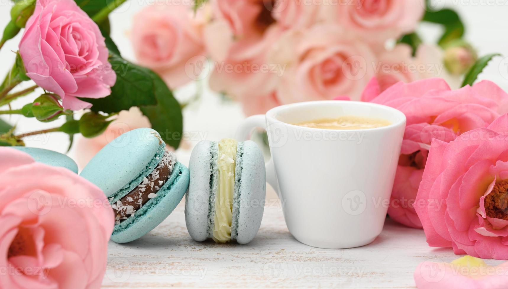 white ceramic cup with coffee and  macarons on a white table, behind a bouquet of pink roses photo
