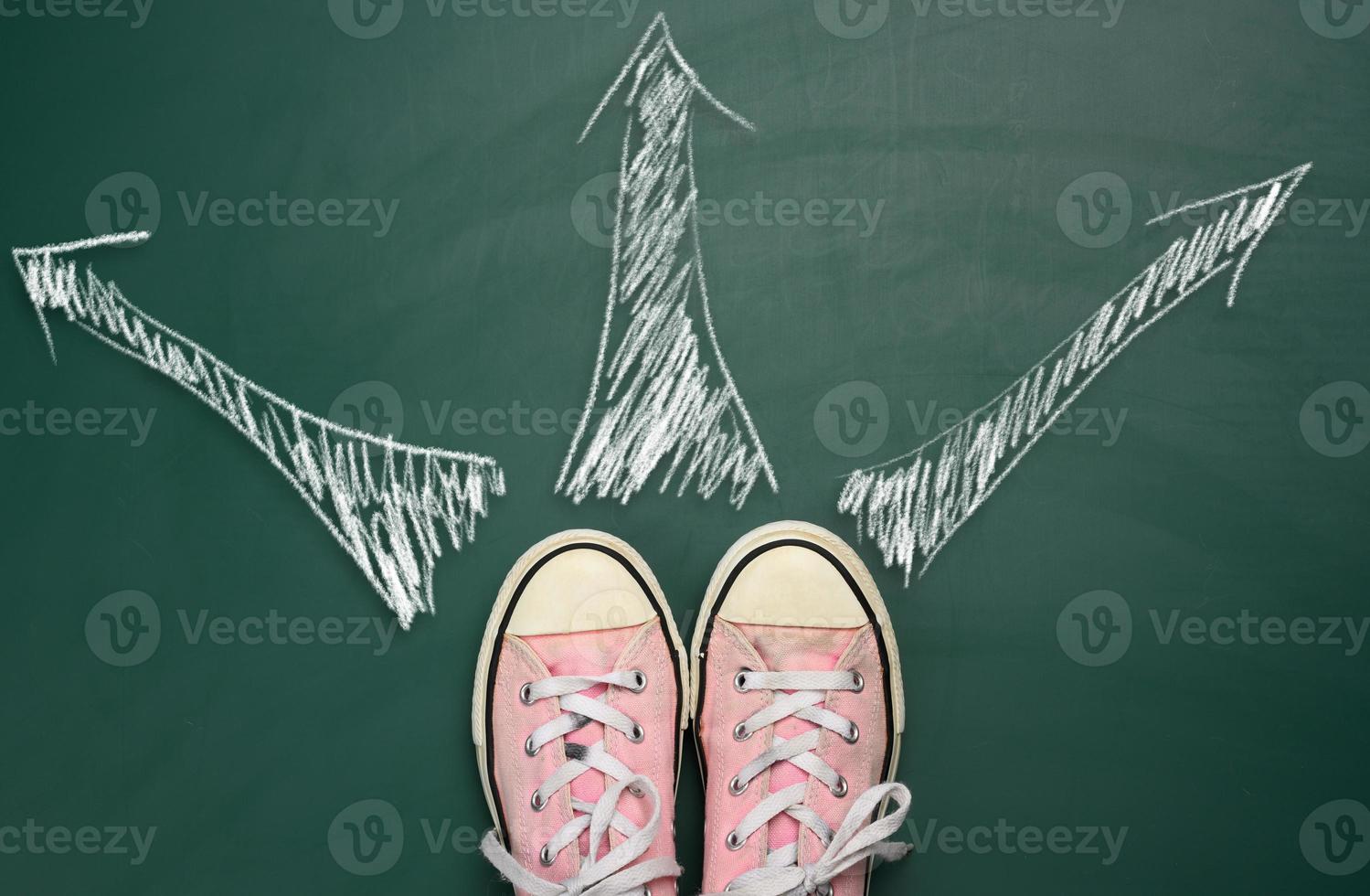 pair of pink sneakers on a green background and arrows drawn in equal directions in white chalk, top view photo