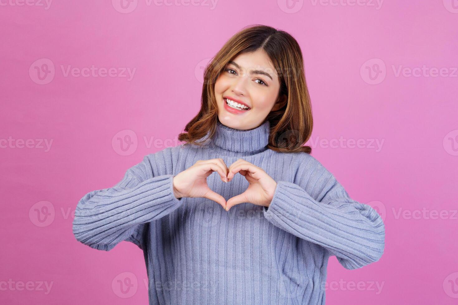 Portrait of Smiling young woman feels happy and romantic shapes heart gesture isolated over pink background photo