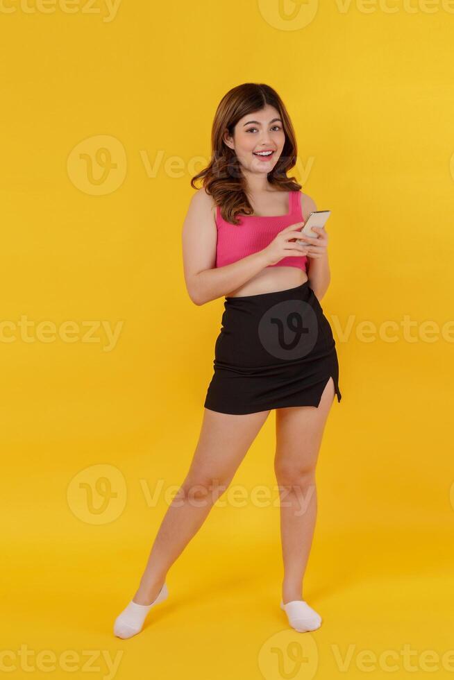 Portrait of Smiling young woman using mobile phone in her hands and looking at camera while standing isolated over yellow background photo