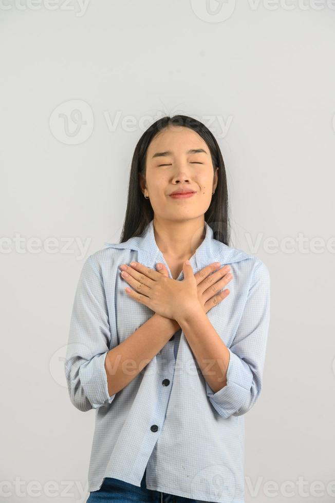 Portrait of Happy young asian woman in blue shirt showing her heart filled with love and holding hands on her breast isolated on white background photo