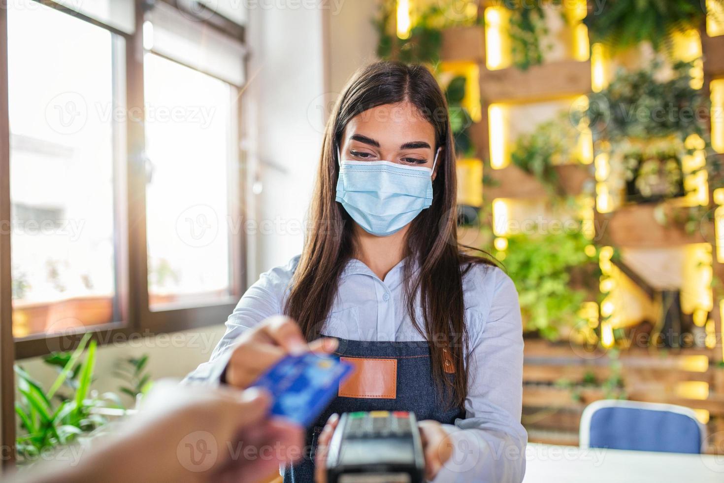 Waitress holding credit card reader machine and wearing protective face mask with client holding credit card. Man hand of customer paying with contactless credit card with NFC technology. photo