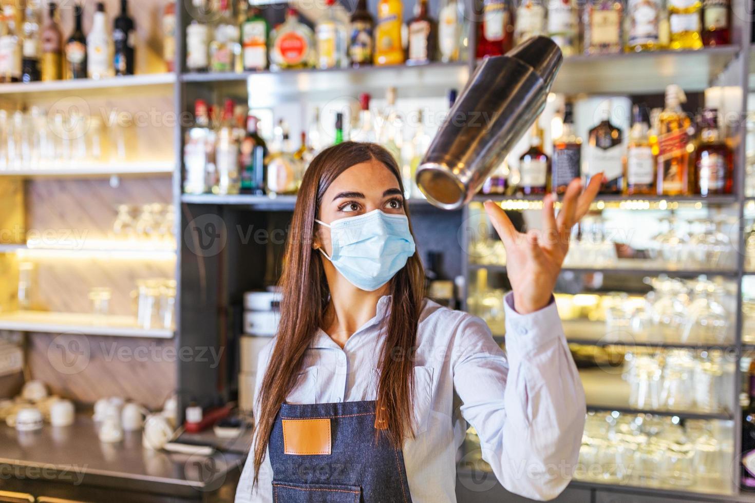 Beautiful female bartender with protective face mask tosses cocktails shaker into the air and arranges a real show for their guests during coronavirus pandemic. New normal photo