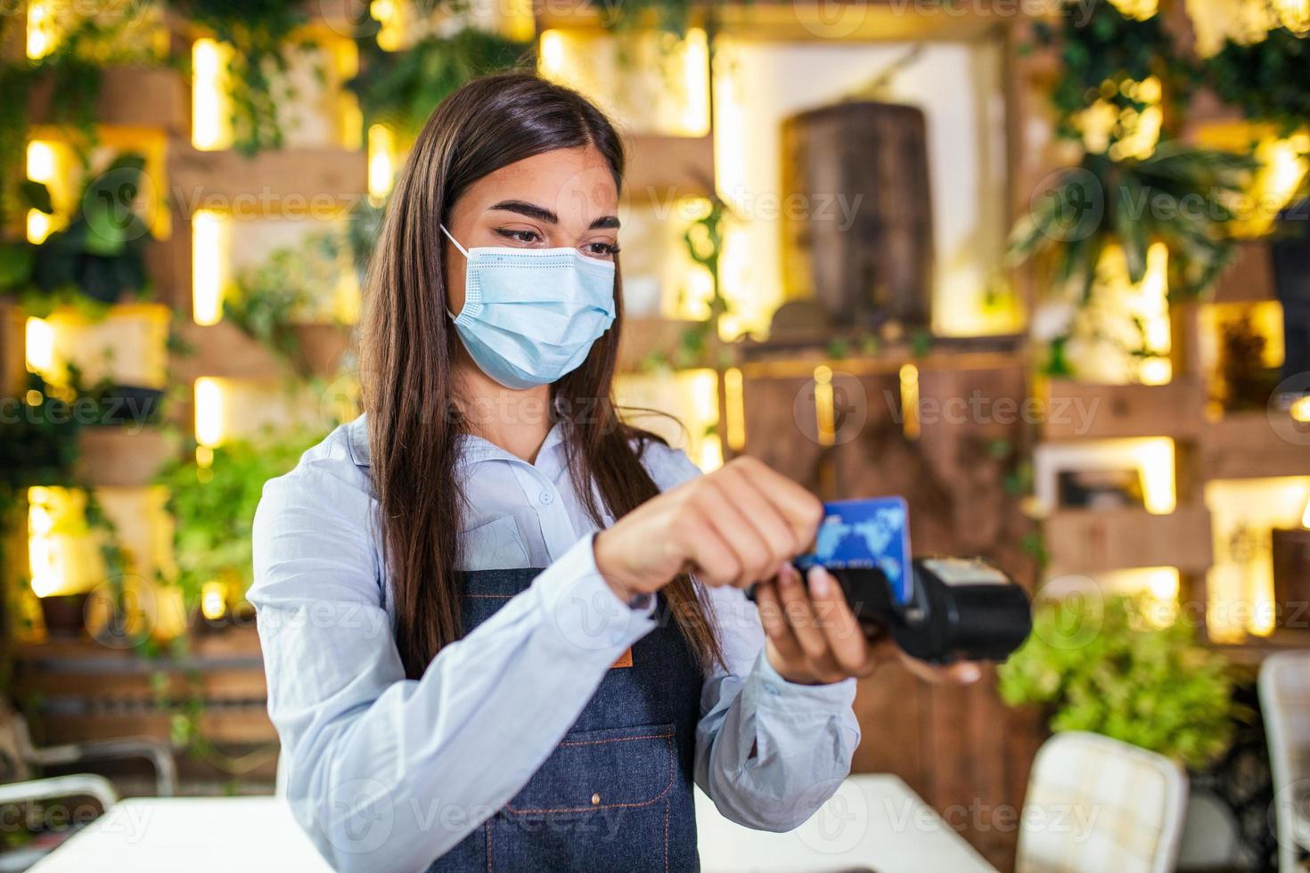 camarera con mascarilla protectora haciendo un pago con tarjeta de crédito con una máquina lectora de tarjetas. pasar la tarjeta de crédito a través de la terminal en el restaurante. mano de la camarera deslizando la tarjeta de débito para pagar la cuenta foto