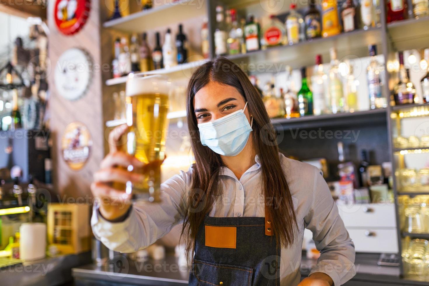 Beautiful female Bartender wearing protective face mask, serving a draft beer during coronavirus pandemic, shelves full of bottles with alcohol on the background photo