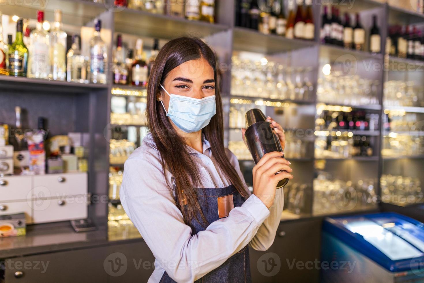 joven trabajadora en el mostrador de la camarera en el bar del restaurante preparando un cóctel con agitador. bella joven detrás de la barra con mascarilla protectora foto