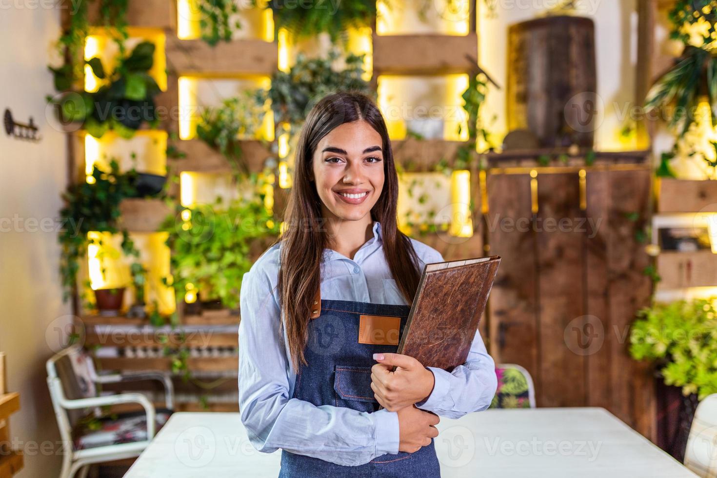 feliz hermosa camarera sonriente con delantal sosteniendo un menú de carpetas en un restaurante, mirando la cámara, de pie en una cafetería acogedora, buen servicio foto