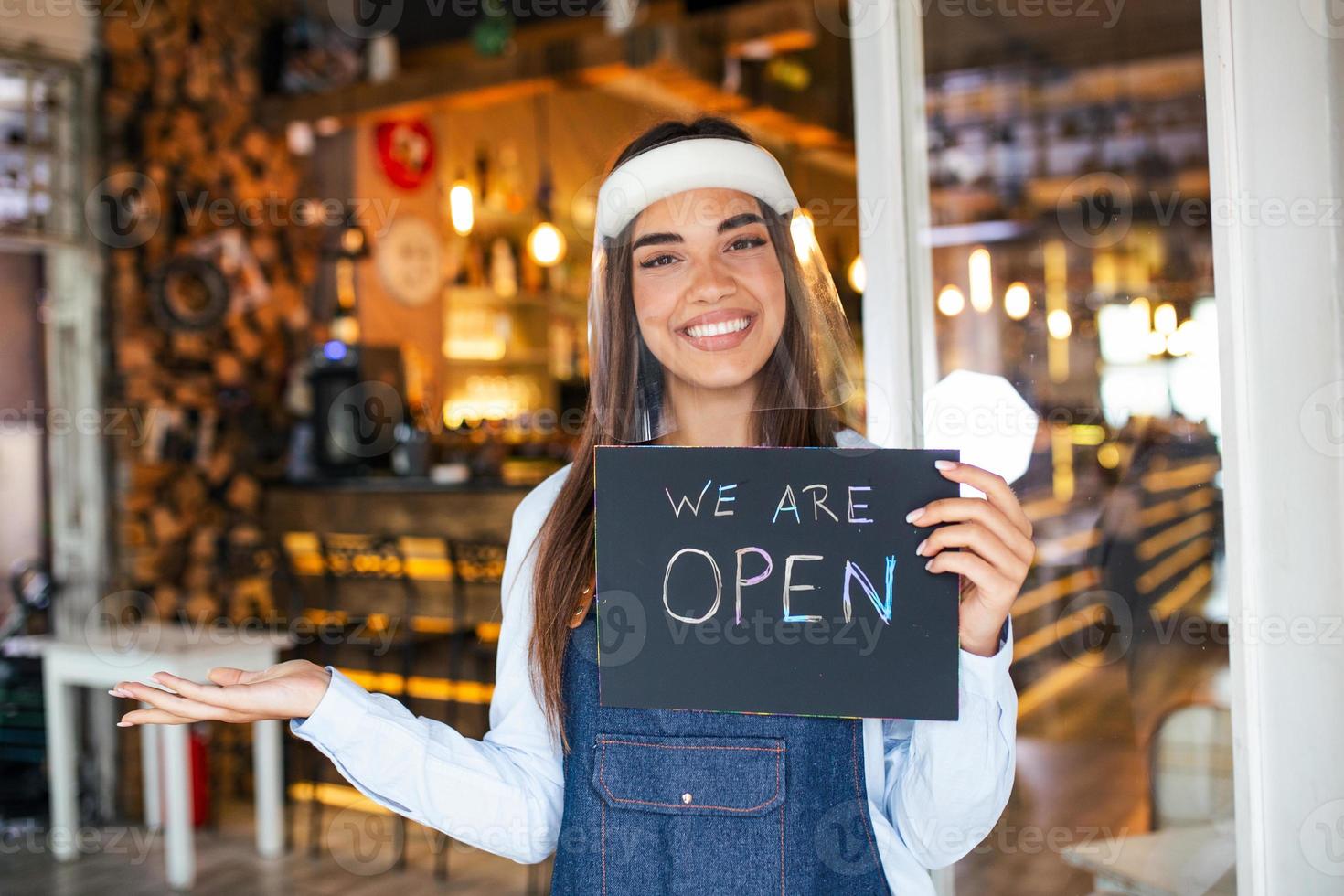 propietario de una pequeña empresa sonriendo mientras sostiene el cartel para la reapertura del lugar después de la cuarentena debido al covid-19. mujer con escudo facial sosteniendo el cartel de que estamos abiertos, apoya los negocios locales. foto