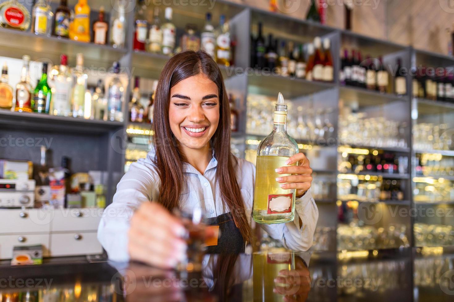 Beautiful female bartender with protective face maskholding shot glass with alcohol during coronavirus pandemic, shelves full of bottles with alcohol on the background photo