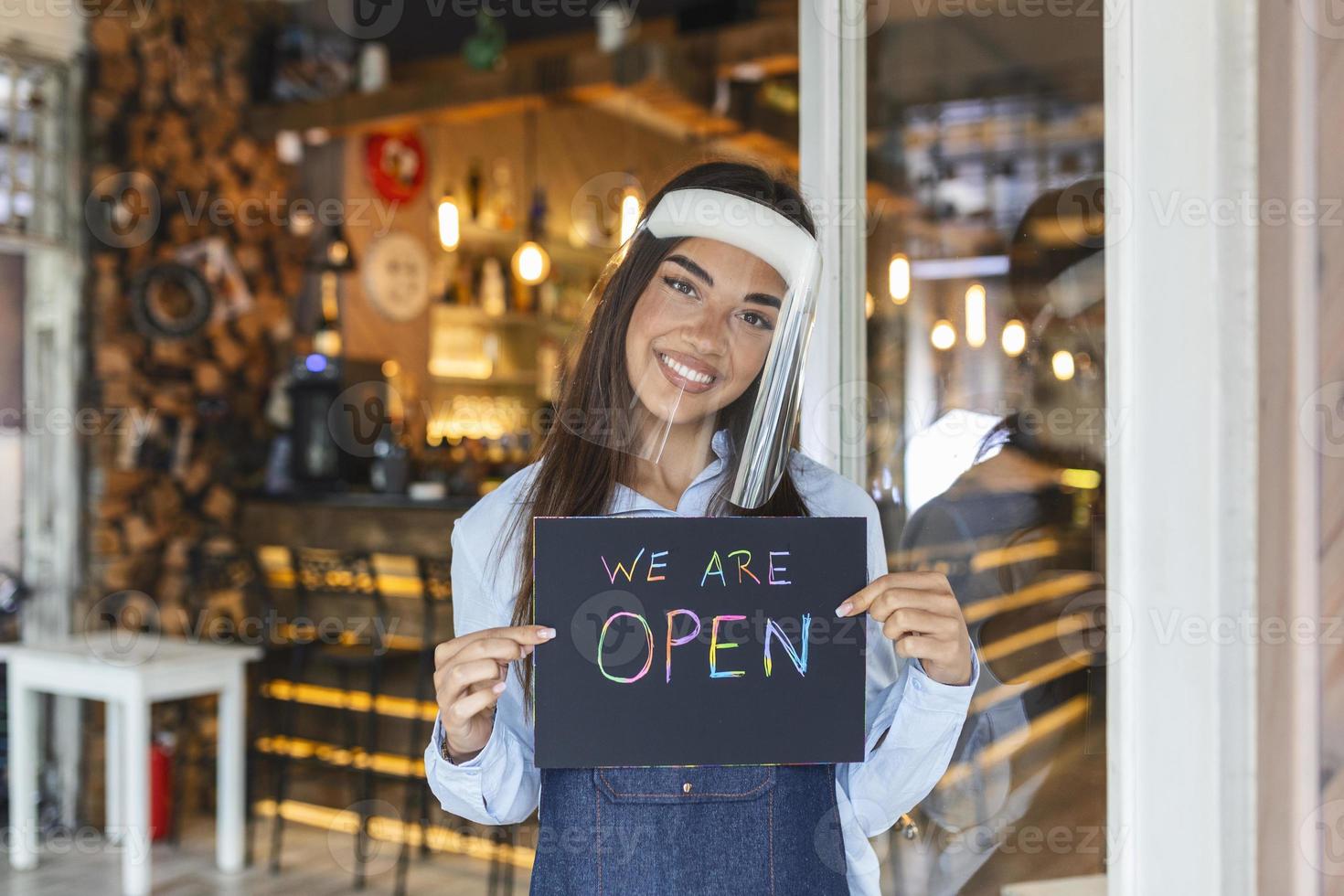 propietario de una pequeña empresa sonriendo mientras sostiene el cartel para la reapertura del lugar después de la cuarentena debido al covid-19. mujer con escudo facial sosteniendo el cartel de que estamos abiertos, apoya los negocios locales. foto