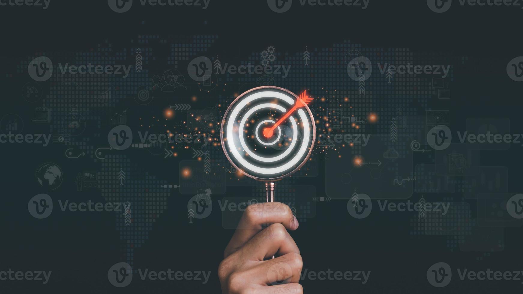 magnifying glass in businessman hand and arrow icon hitting center of dartboard target,Setting business goals and focused concepts, Organizational growth and objectives, marketing strategy planning photo