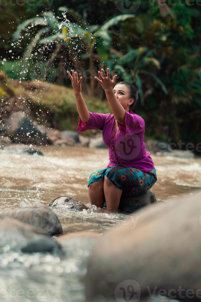 mujer asiática jugando con agua sucia de un río sucio mientras usa un vestido morado y una falda verde foto