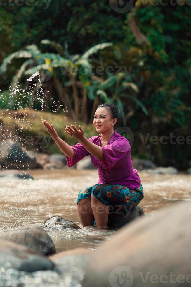 mujer asiática jugando con agua sucia de un río sucio mientras usa un vestido morado y una falda verde foto