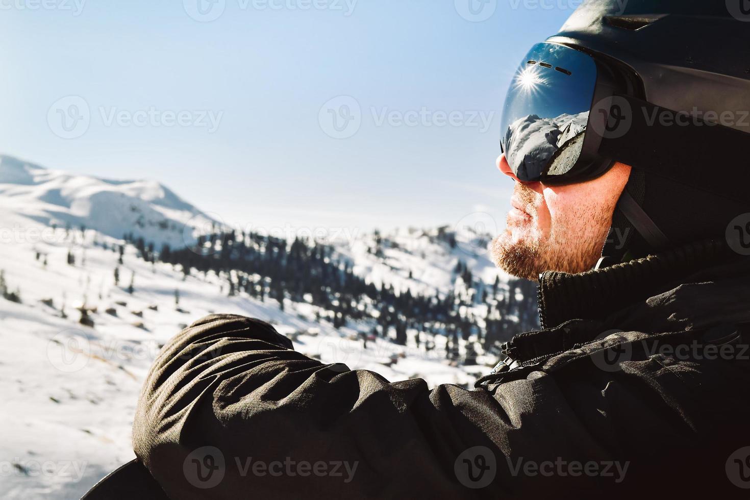 Close up of the ski goggles of caucasian man with the reflection of snowed mountains and sunburst. A mountain range reflected in the ski mask. Portrait of man at the ski resort photo