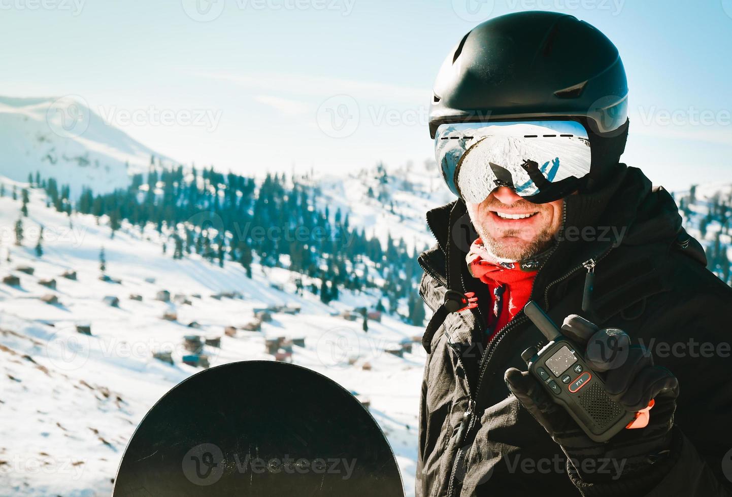 hombre caucásico con chaqueta negra y gafas con casco de esquí en la cabeza sostiene un walkie-talkie en la mano contra el fondo de la nieve blanca foto