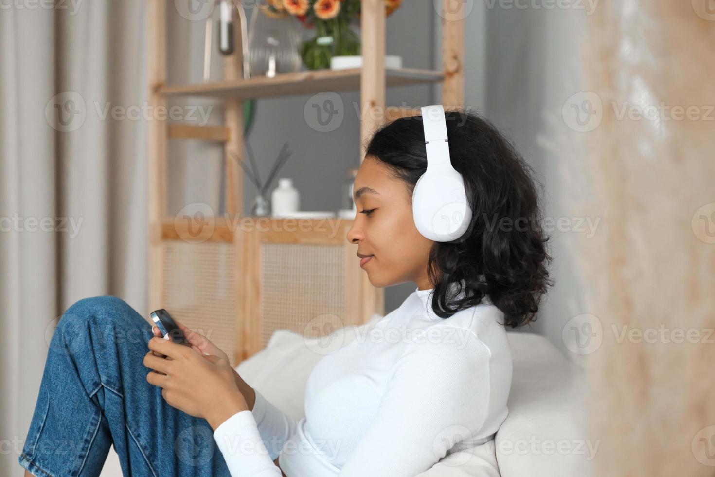 Side view portrait of a happy african amercian girl listening music,podcast or lecture on sofa at home with phone in her hands. Online application and modern technologies concept. photo