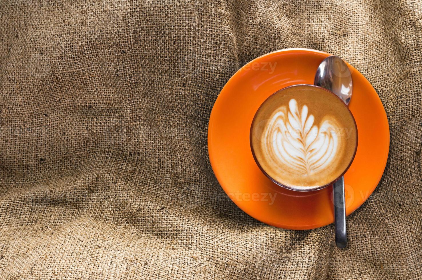 Top view hot latte in orange cup with floral pattern in foam on burlap background, brown sackcloth, vintage beige fabric texture photo