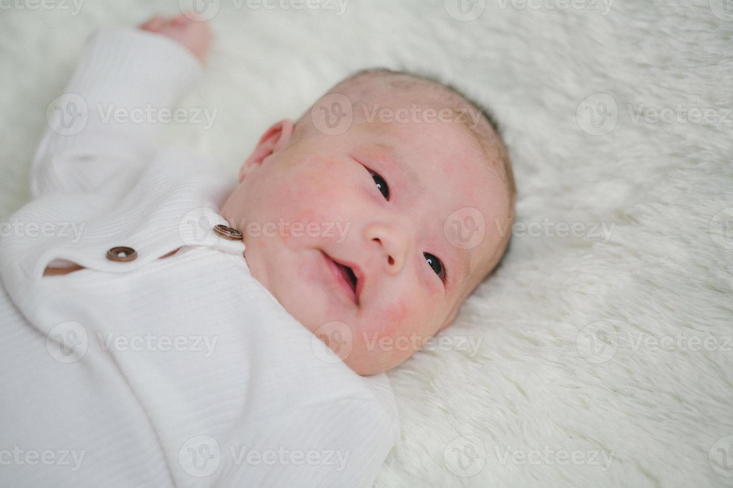 Closeup cute newborn baby in white bodysuit lying down alone on bed. Adorable infant rests on white bedsheets, staring at camera looking peaceful. Infancy, healthcare and paediatrics, babyhood concept photo