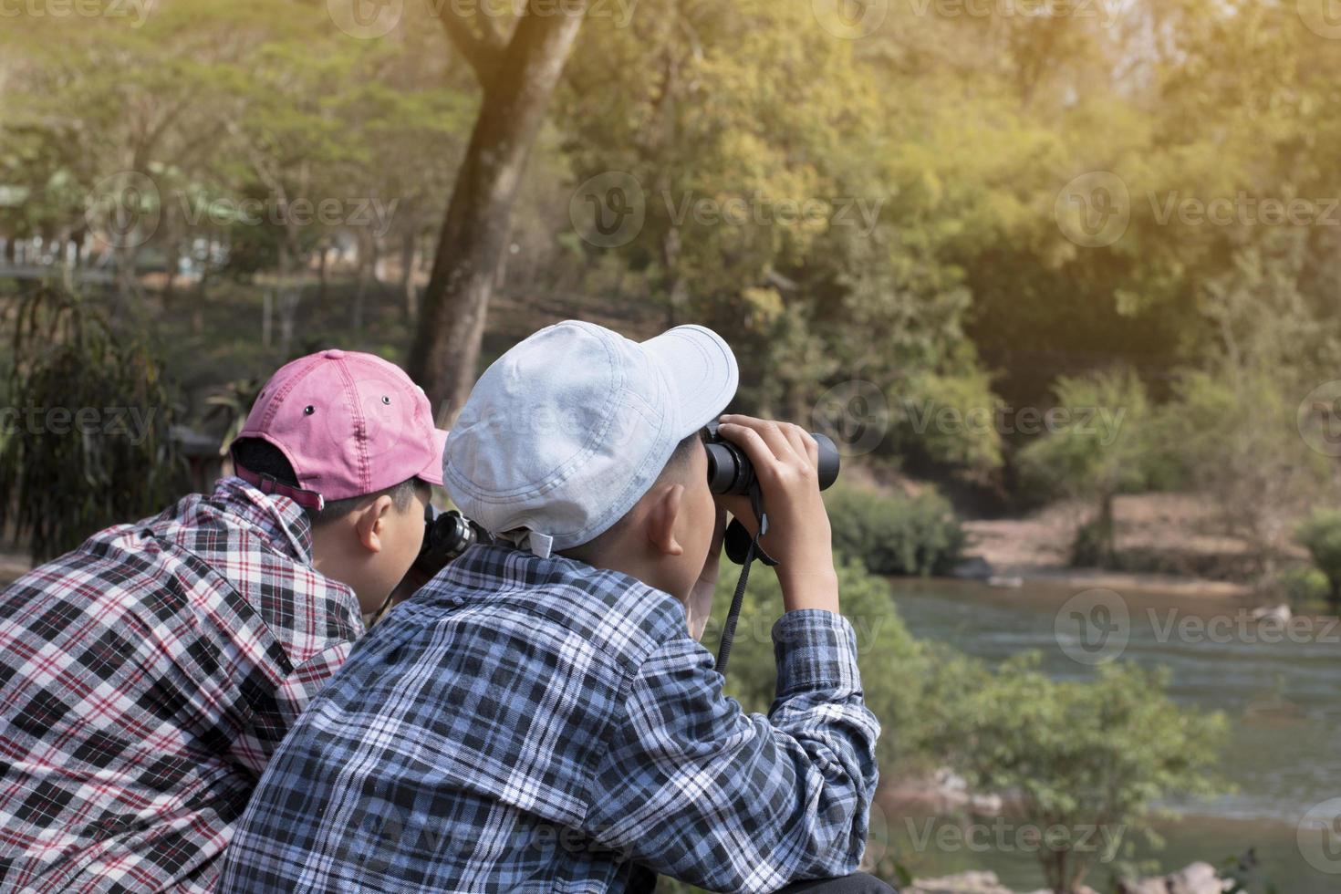 Asian boys using binoculars to watch birds on trees and fish in river in local national park during summer camp, idea for learning creatures and wildlife animals and insects outside the classroom. photo