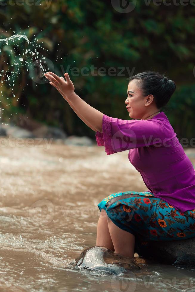 Happy Asian woman playing with the water near the river while sitting on the rock and wearing a purple dress photo