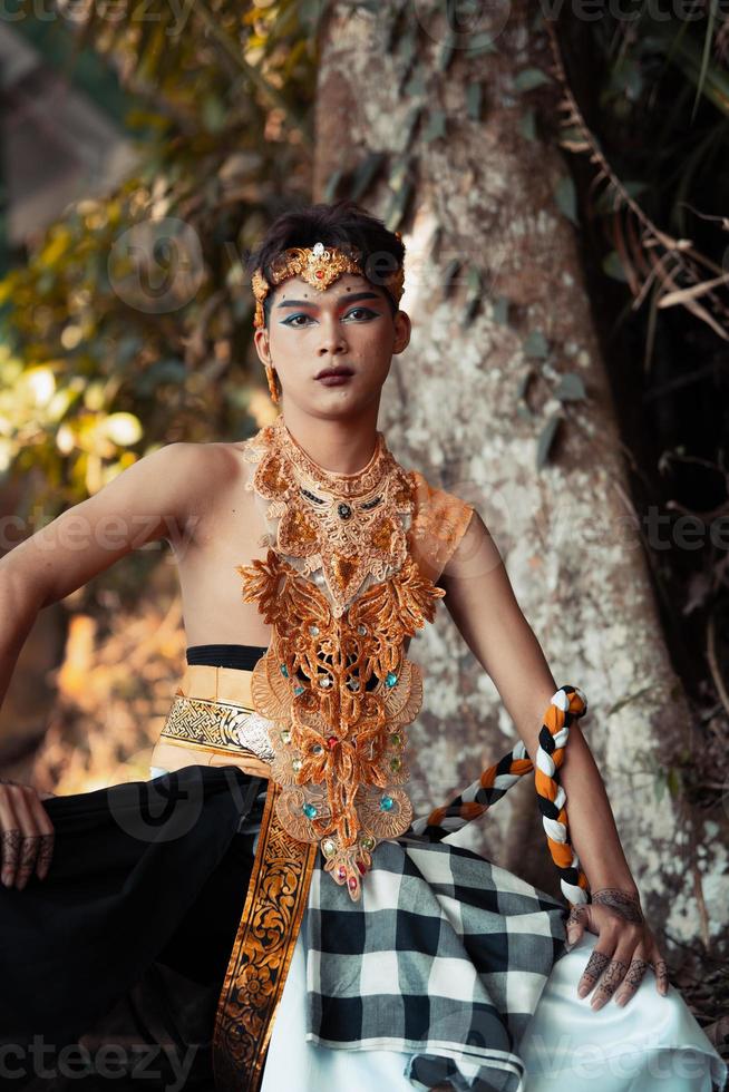 Cute Balinese man sitting in a gold costume shirtless with makeup on his face photo