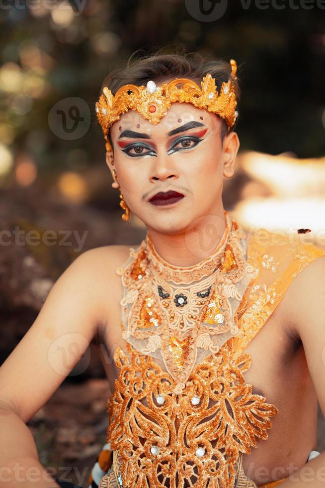 hombre balinés con sombra de ojos y lápiz labial negro en la cara mientras usa un traje dorado y una corona foto