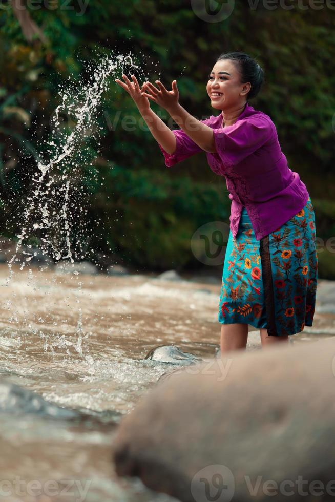 A beautiful Asian woman washing her hand and playing with the water while standing near the river in a traditional purple dress and green skirt photo