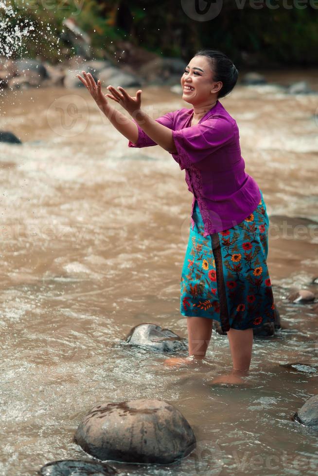 una hermosa mujer asiática lavándose la mano y jugando con el agua mientras está de pie cerca del río con un vestido morado tradicional y una falda verde foto