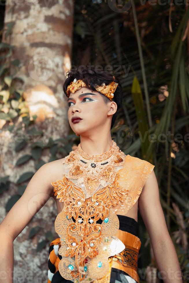 A Balinese man with a golden costume and makeup on his face is standing in front of the coconut tree photo