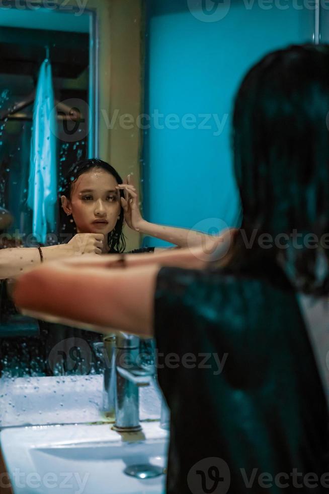 Indonesian woman staring the mirror of herself wearing a black dress with wet body and wet hair photo