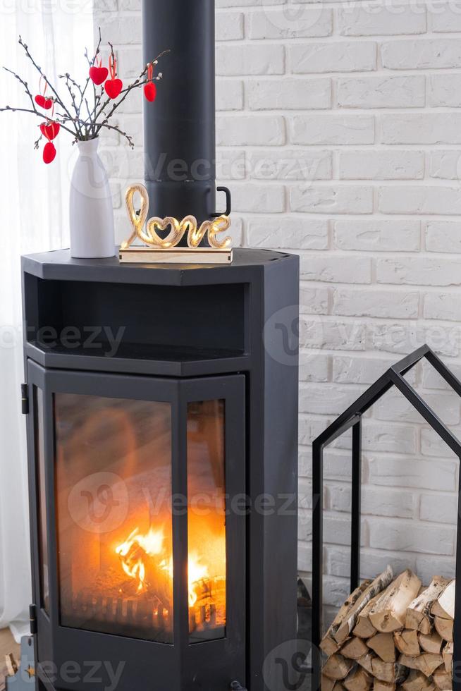 Valentine decor near fireplace stove with fire and firewood. Cozy home hearth in interior with potted plans, valentines day in family love nest. photo