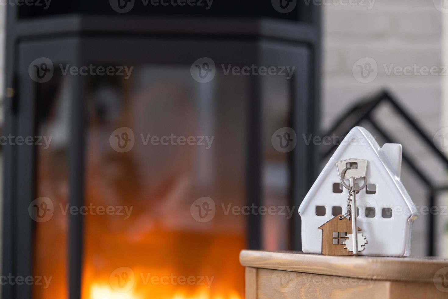 Key to house with keychain against background of fireplace stove with fire and firewood. Cozy home hearth. Building, design, project, moving to new house, mortgage, rent and purchase real estate photo