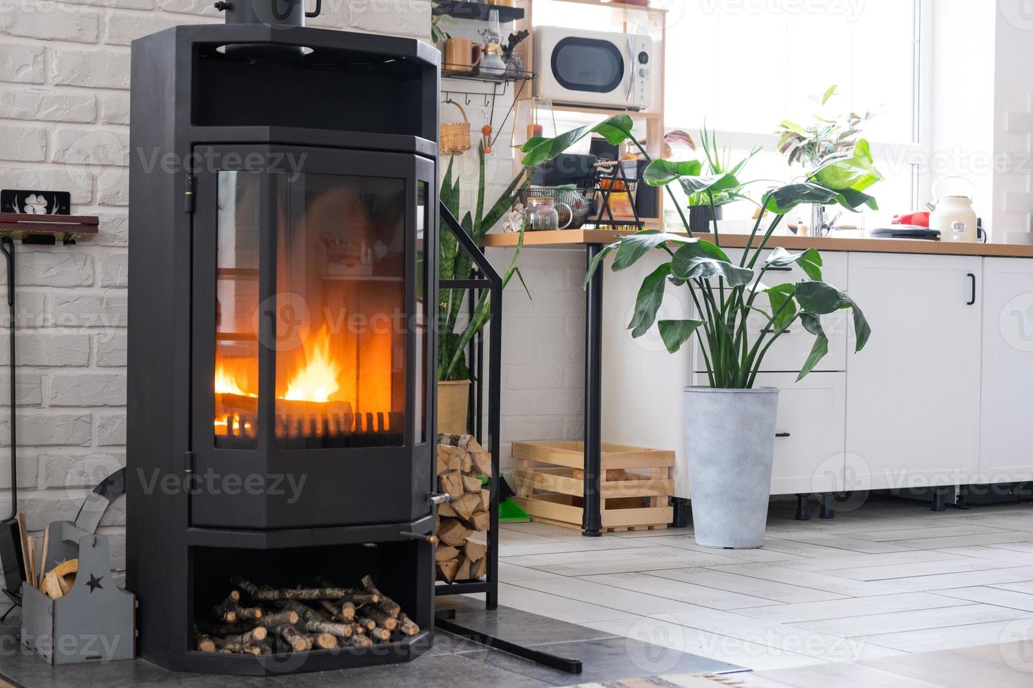 Estufa de chimenea de acero negro con fuego y leña cerca de la cocina en casa de pueblo. acogedor hogar hogar en el interior con plantas en macetas strelitzia reginae foto