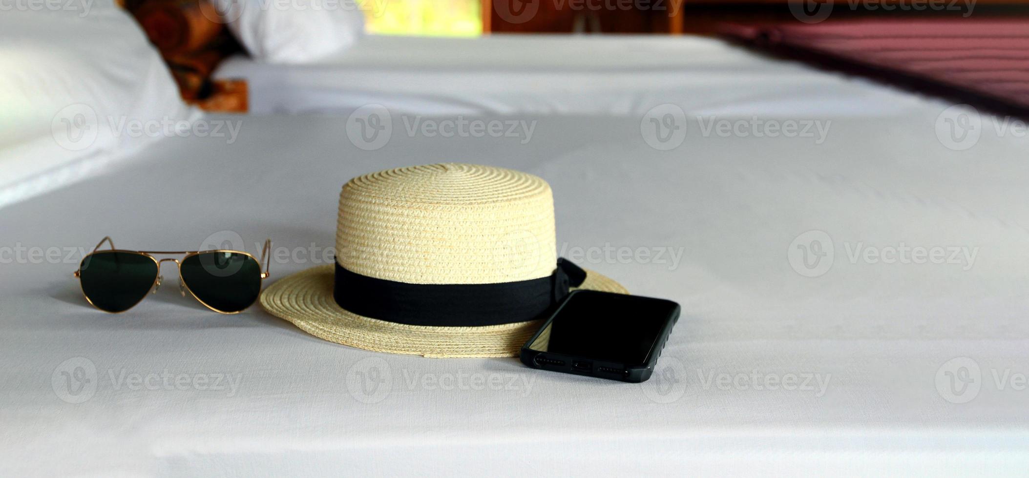 tiempo de vacaciones el fin de semana con gafas de sol negras, sombrero de paja vintage blanco y teléfono inteligente en una cama blanca con espacio para copiar. concepto de viajes, accesorios, moda y tiempo de descanso foto