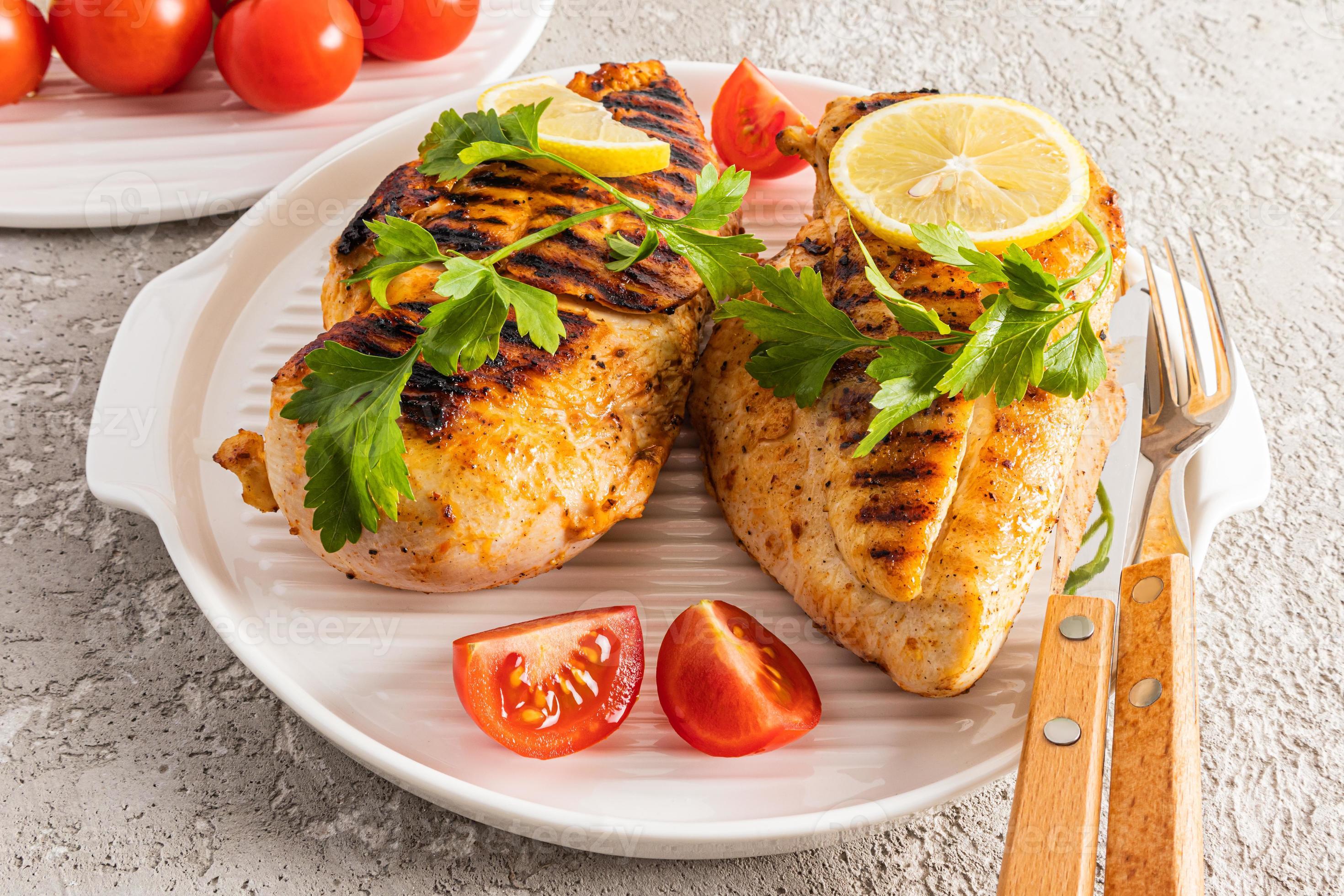 two grilled chicken breasts on a white serving tray with parsley, lemon, tomato slices. closeup