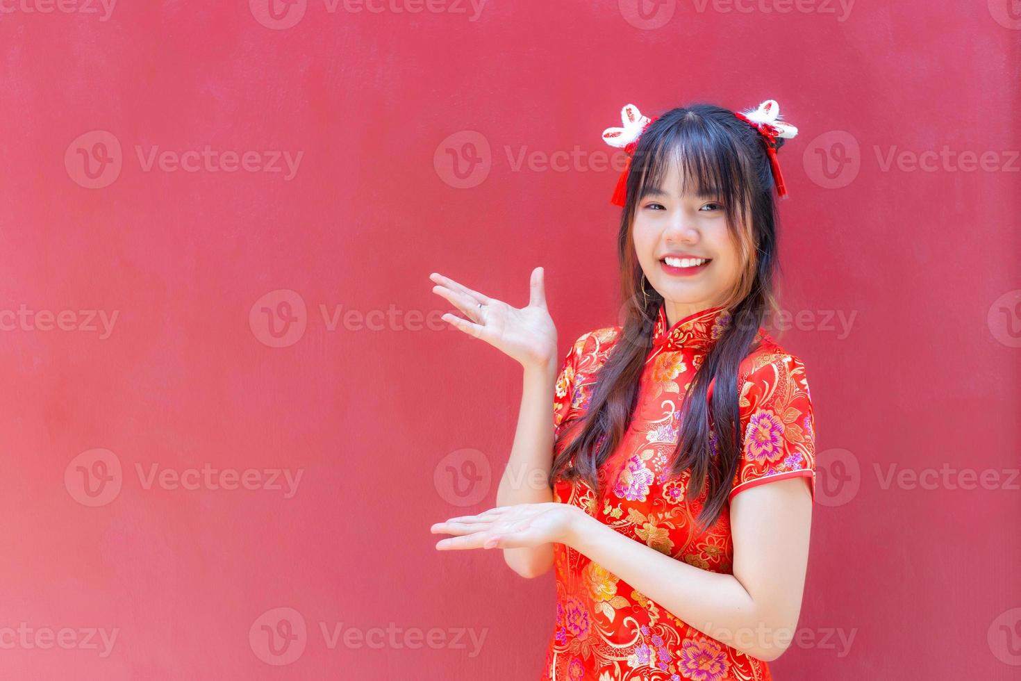 Beautiful and pretty young Asian girl with long hair who wears a red Cheongsam dress in Chinese new year theme while she shows her hand to present something on red background. photo