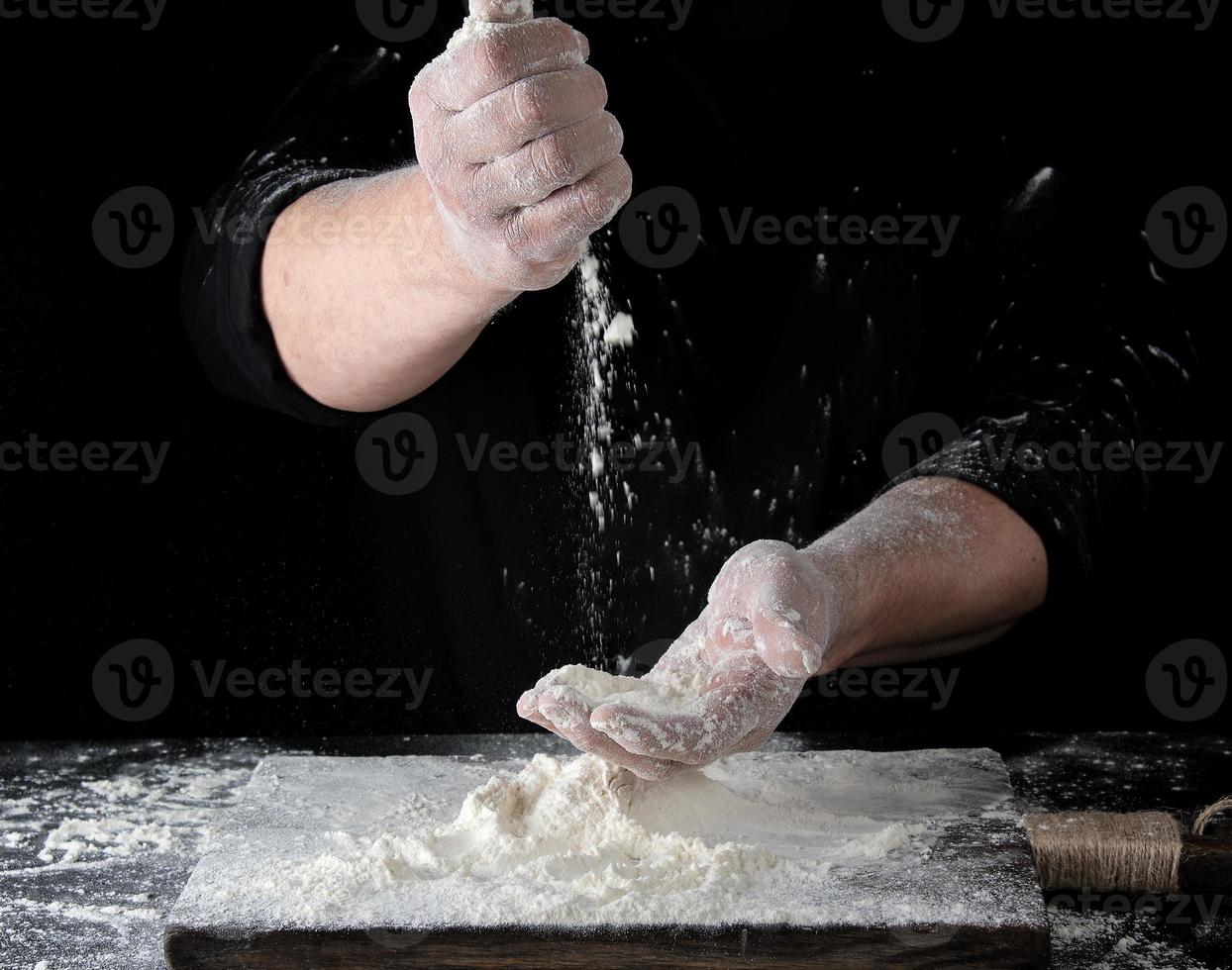 chef en uniforme negro tamiza a través de sus dedos harina de trigo blanco foto