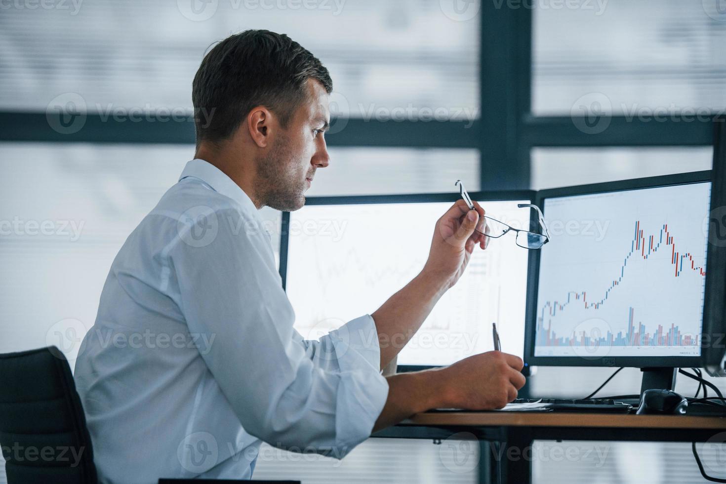 Side view. Young businessman in formal clothes is in office with multiple screens. Conception of exchange and money photo