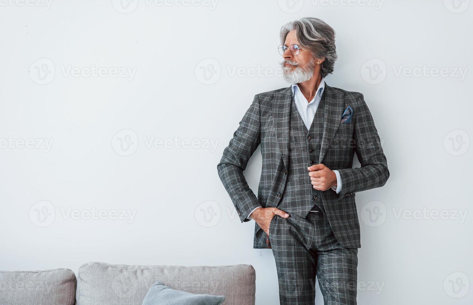 In elegant clothes. Standing against white wall. Senior stylish modern man with grey hair and beard indoors photo