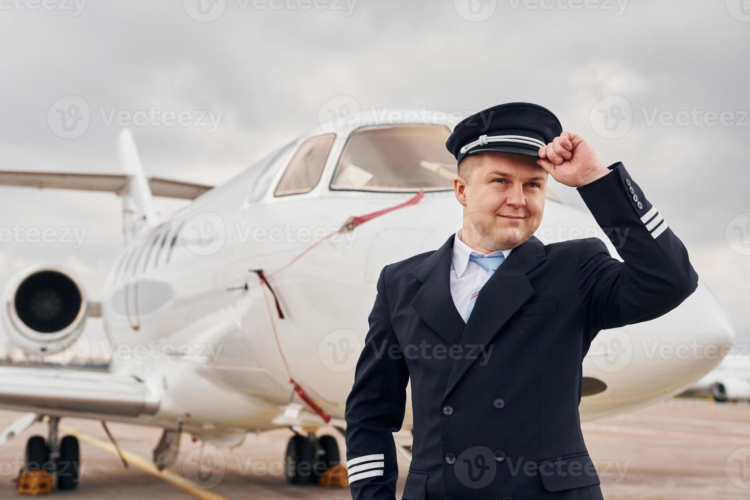 Posing for a camera. Experienced pilot in uniform standing outside near plane photo