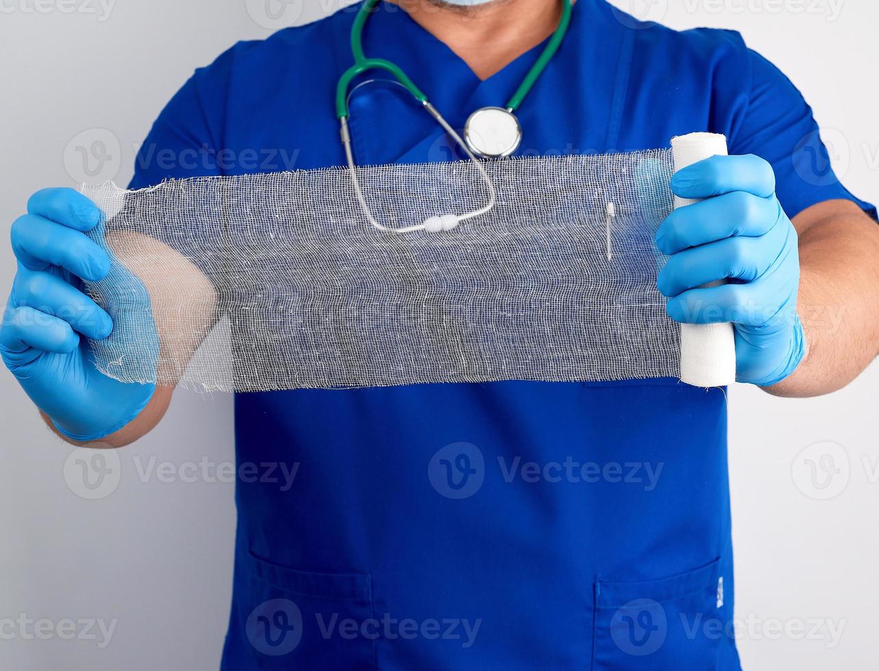 médico con uniforme azul y guantes de látex sostiene un rollo de vendaje blanco para vendar heridas de gasa foto