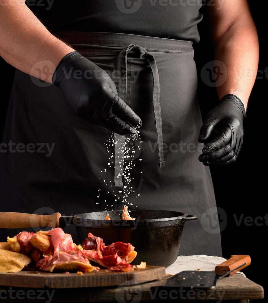 chef con uniforme negro y guantes de látex rociados con carne de pollo cruda con sal blanca en una sartén negra de hierro fundido foto
