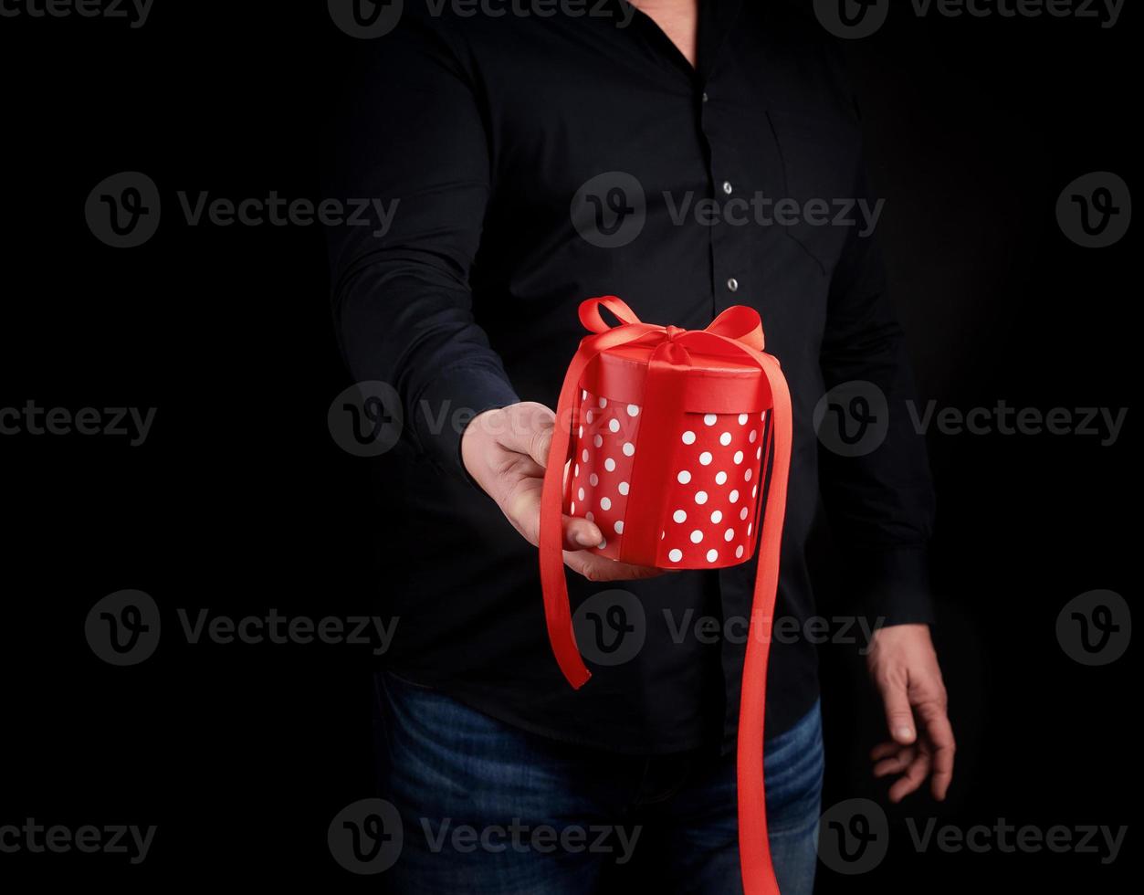 adult man in a black shirt holds in his hand a round red box of polka dots with a bow on a black background photo