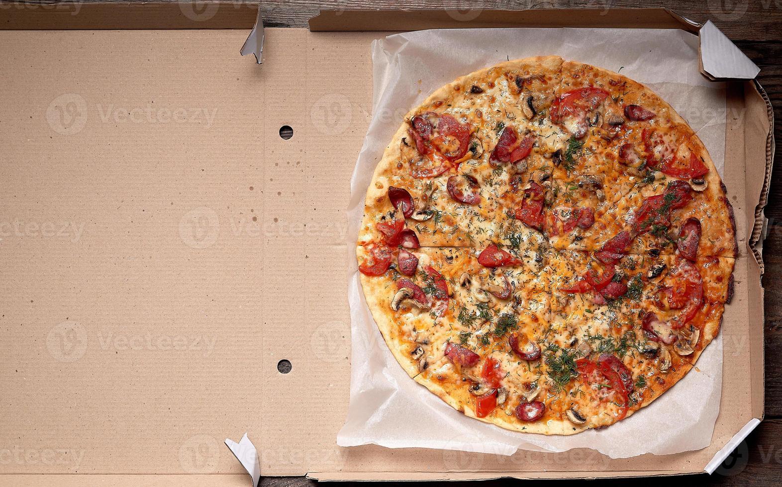 baked round pizza with smoked sausages, mushrooms, tomatoes, cheese and dill in an open cardboard box photo