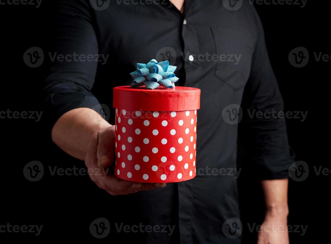 adult man in a black shirt holds in his hand a round red box of polka dots with a blue bow photo
