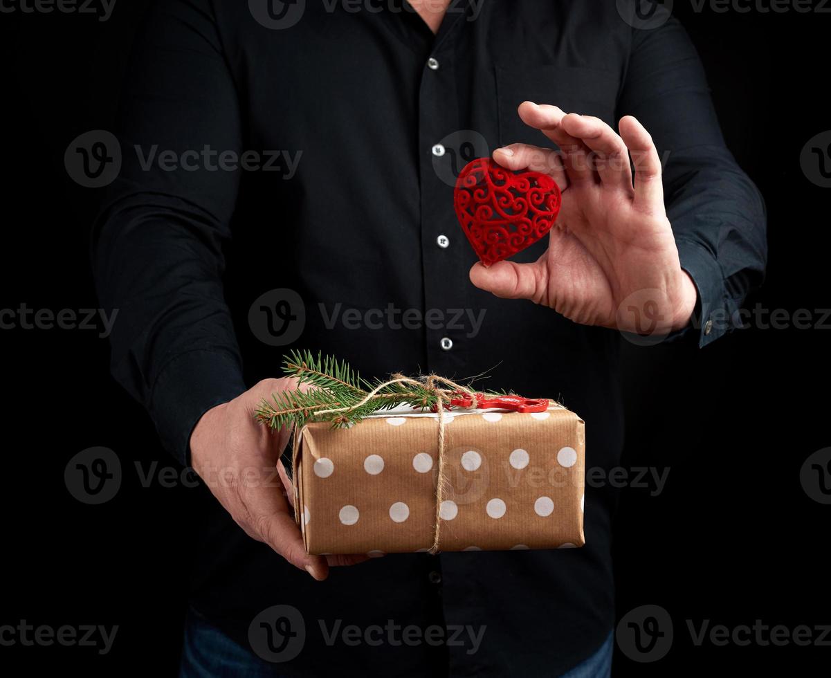 adult man is standing on a dark background wearing a black shirt and holding a wrapped gift in brown paper photo