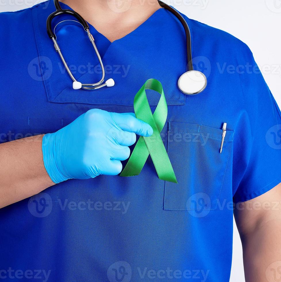 doctor in blue uniform and latex gloves holds a green ribbon as a symbol of early research and disease control photo