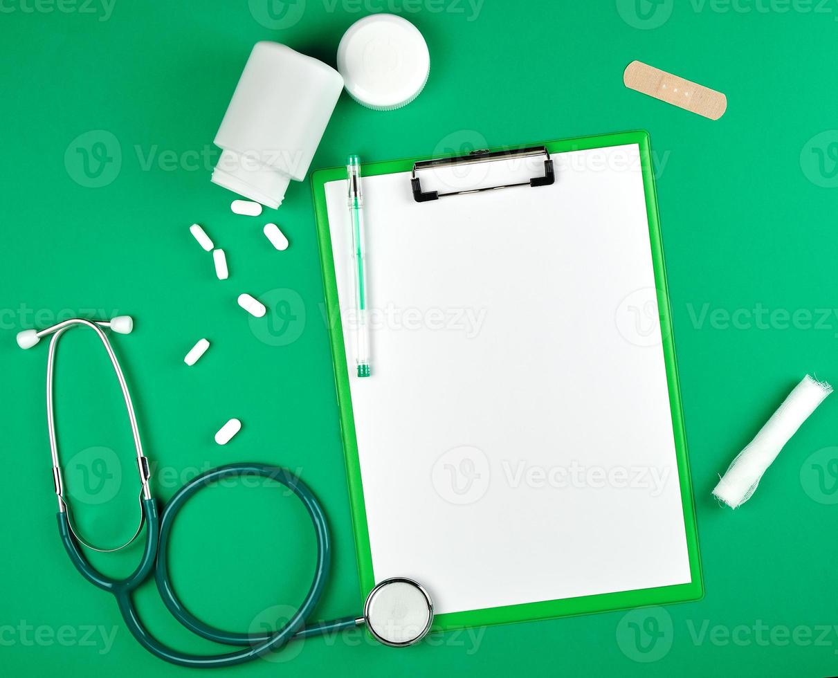 paper holder with empty white sheets, medical stethoscope, pills on a green background photo