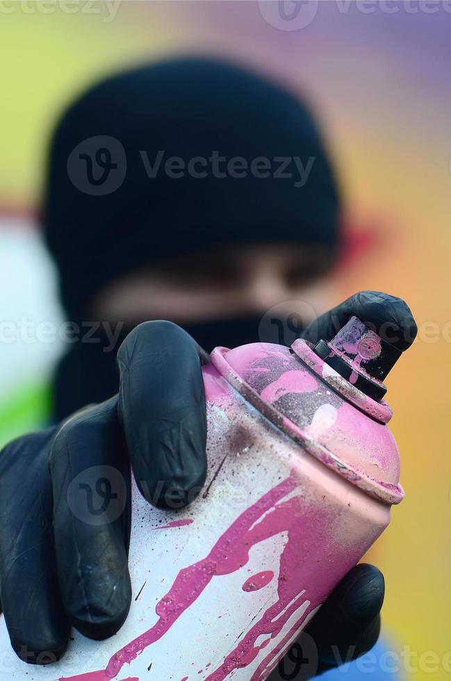 A young graffiti artist in a blue jacket and black mask is holding a can of paint in front of him against a background of colored graffiti drawing. Street art and vandalism concept photo